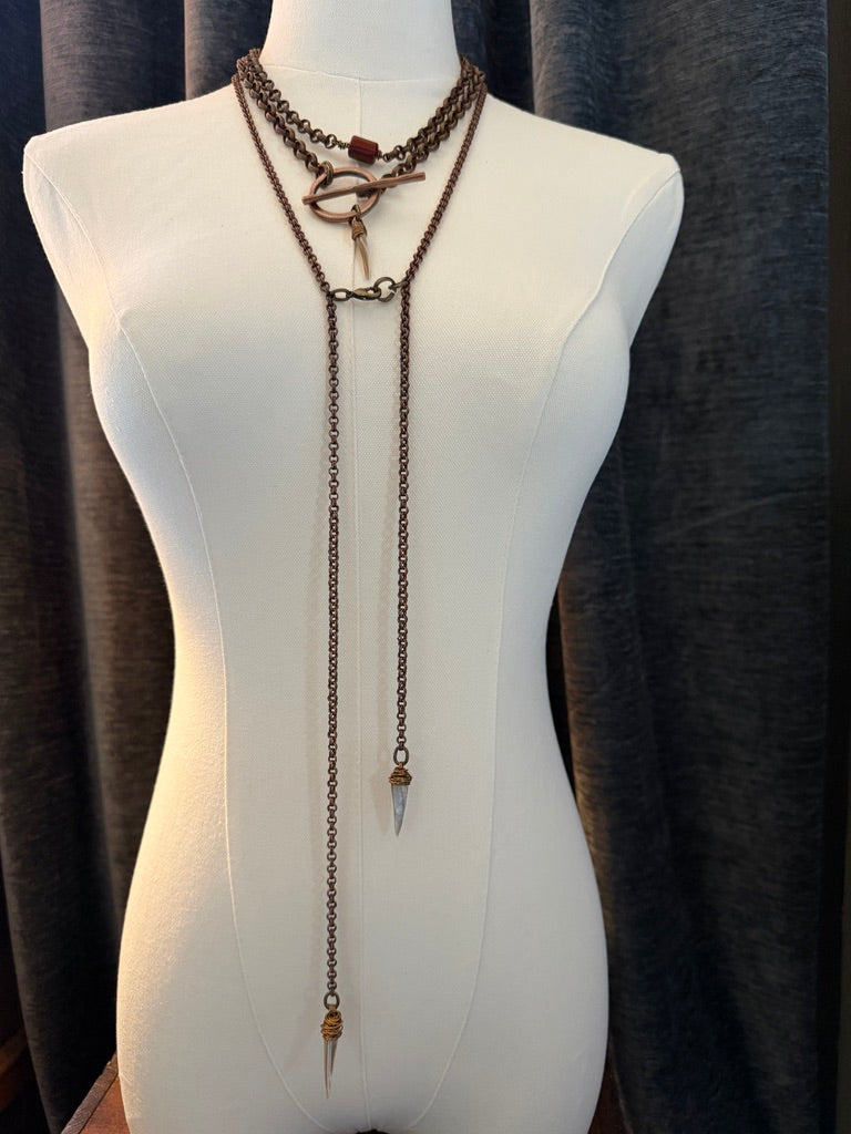 A long layered necklace displayed on a white mannequin torso, featuring dark metal chains, a toggle clasp, and a dangling spear-shaped pendant, set against a dark curtain background.
