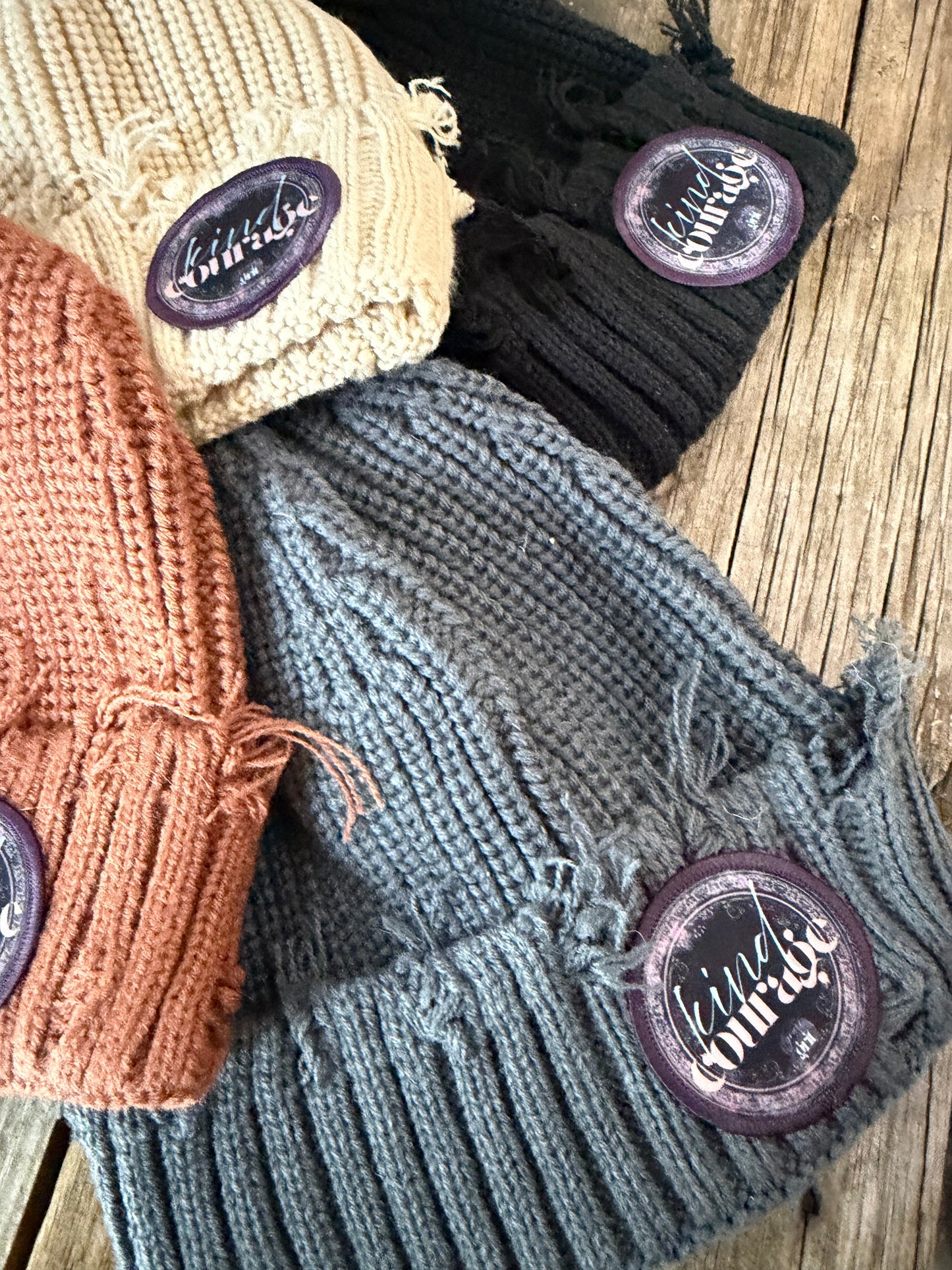 Four knitted hats in black, gray, beige, and brown with branded buttons on a wooden surface.