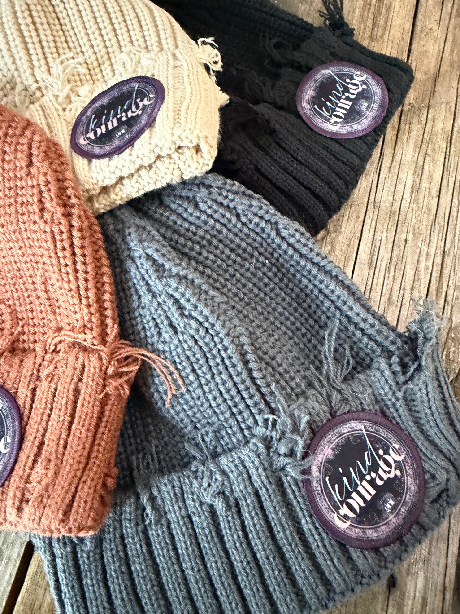 Four knitted hats in black, gray, beige, and brown with branded buttons on a wooden surface.