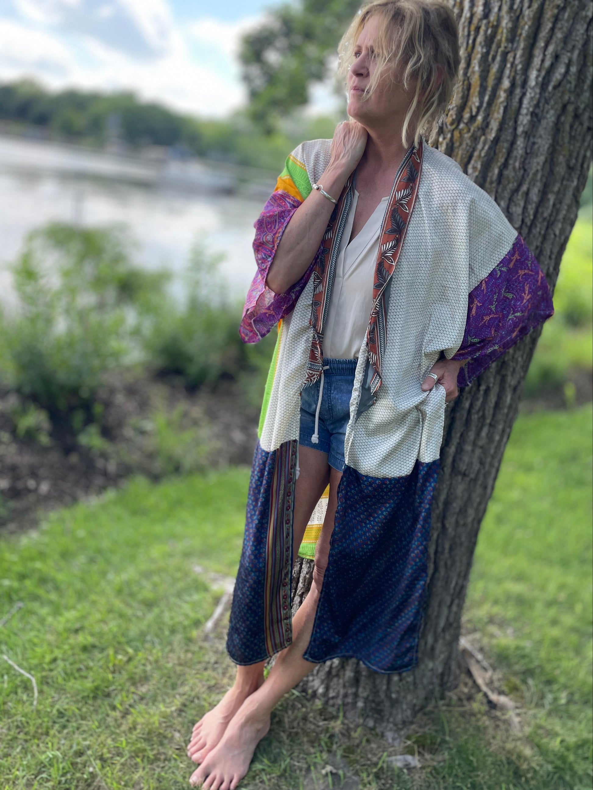 Person leaning against a tree by a body of water wearing a colorful kimono-style garment.