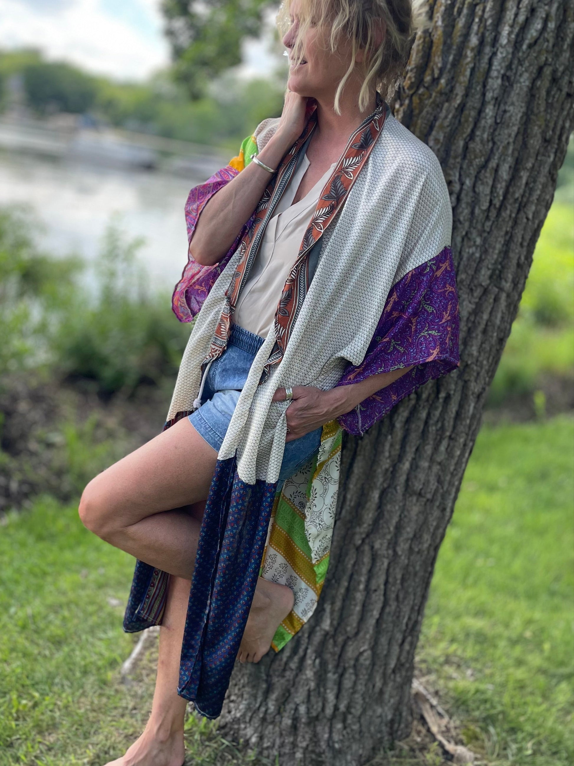 Woman sitting against a tree by a body of water wearing a colorful kimono.