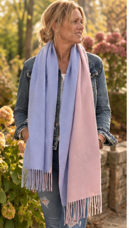 A woman outdoors wearing a long, soft woven scarf in two pastel shades: lavender blue on one side and dusty pink on the other. She is dressed in a denim jacket and jeans, standing in a garden with blurred flowers and foliage in the background.
