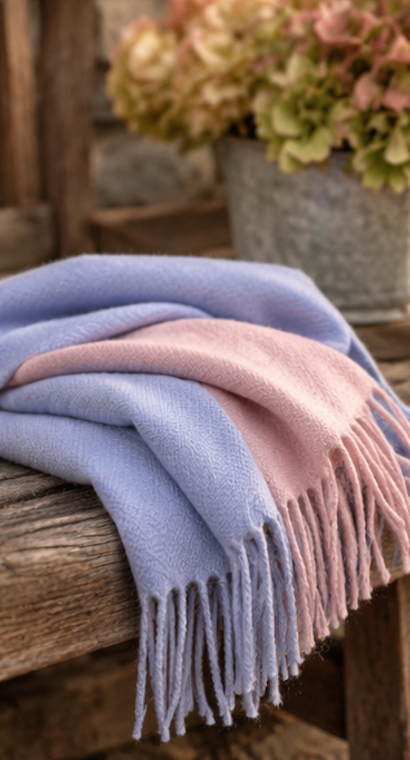 A soft woven throw blanket draped over a rustic wooden bench, featuring a two-tone design in light periwinkle blue and dusty pink with fringed edges. A blurred metal planter with dried flowers sits in the background.