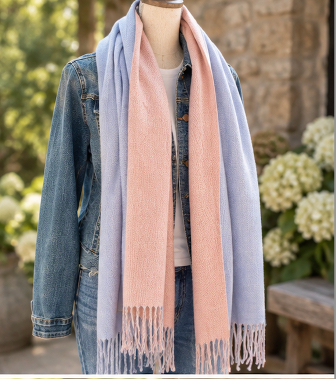 A long, lightweight scarf in pastel light blue and soft peach-pink with fringed ends, draped around a mannequin wearing a denim jacket and jeans, set against a blurred outdoor garden background.