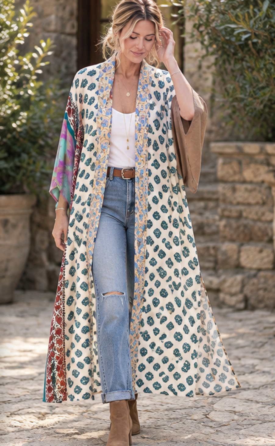 A woman stands outdoors wearing a long cream open-front duster-style cardigan with a blue and gold geometric print, layered over a white tank top and light blue jeans. She has a tan shawl draped over one shoulder and brown ankle boots.