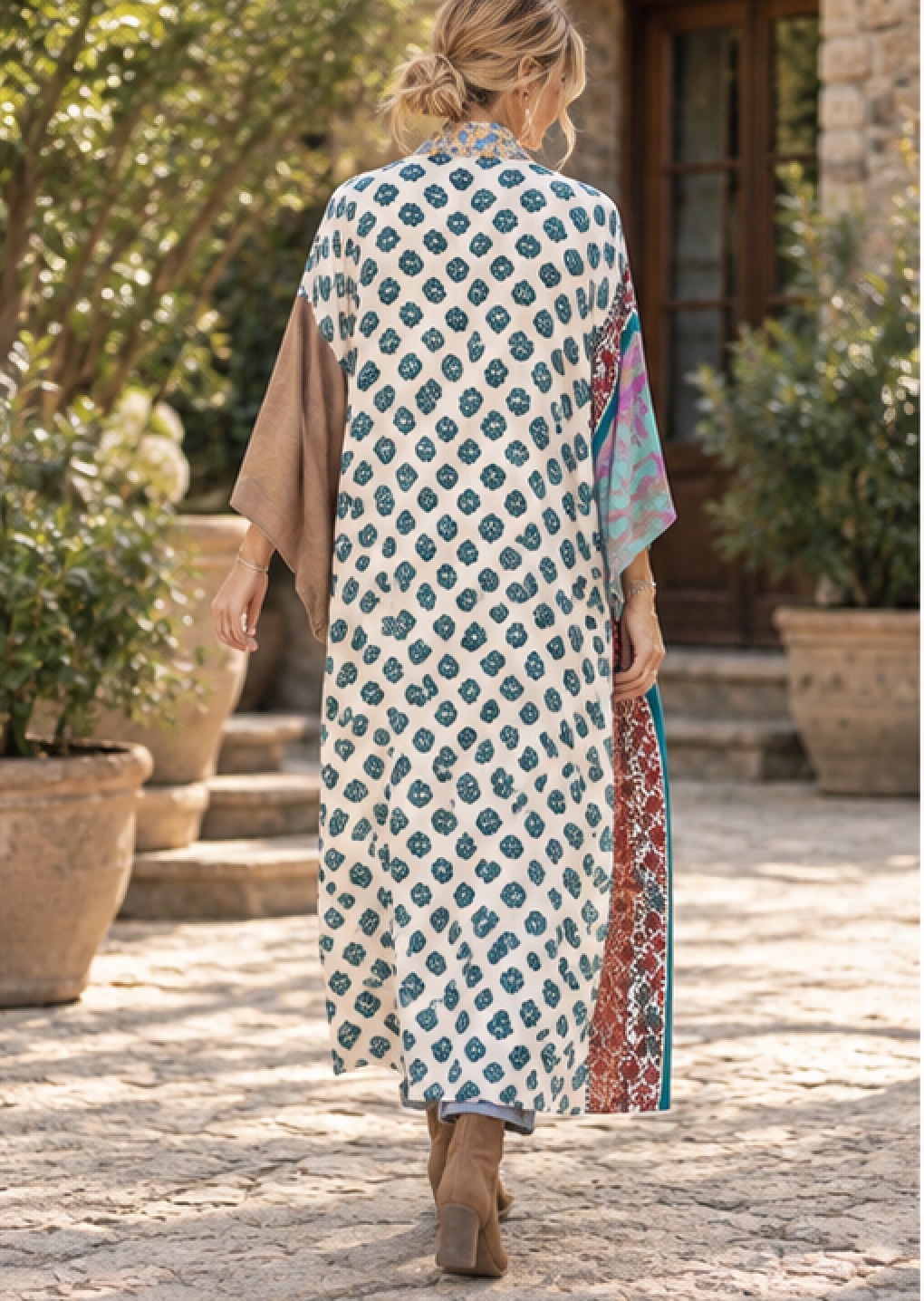 A woman viewed from behind wearing a long white kaftan dress with teal blue floral block prints and brown draped sleeves, walking on a stone path in a garden setting.