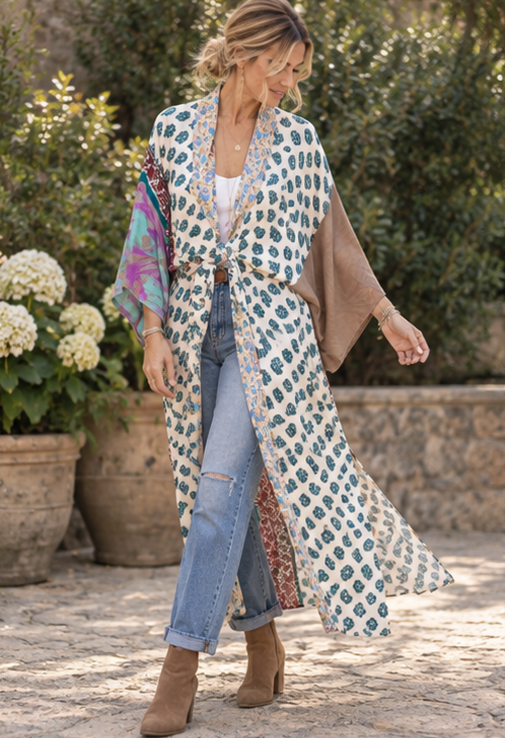 A woman wearing a long cream kimono-style cardigan with a blue and green floral print, styled with light blue jeans, a white top, and brown ankle boots, standing outdoors in front of potted plants.