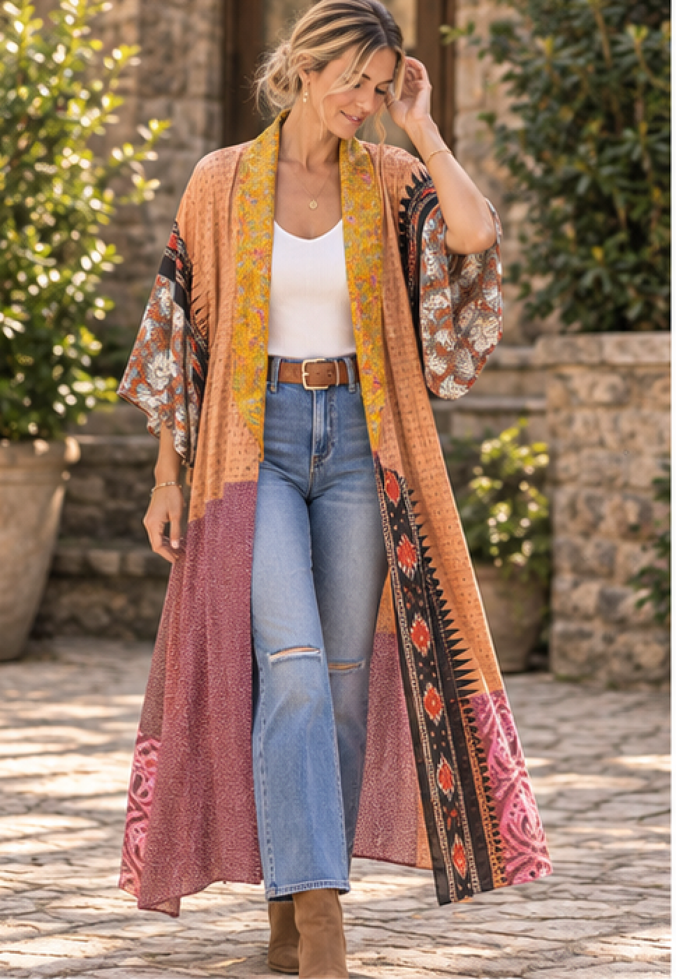 A woman stands outdoors wearing a long, open-front bohemian-style cardigan with wide patterned sleeves and a multicolor geometric border. The cardigan features a warm orange and yellow upper section, a pink lower section, and a white tank top, blue jeans, and brown boots underneath.