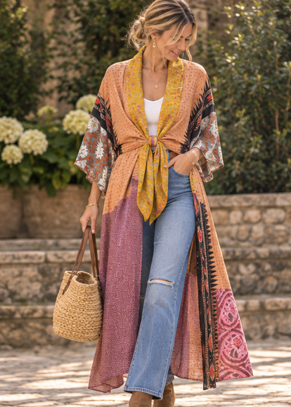 A woman wearing a long, open-front kimono-style cover-up with a tie waist, featuring a multicolor patchwork pattern in mustard yellow, dusty pink, orange, and brown, styled with light blue jeans and a straw bag in an outdoor garden setting.