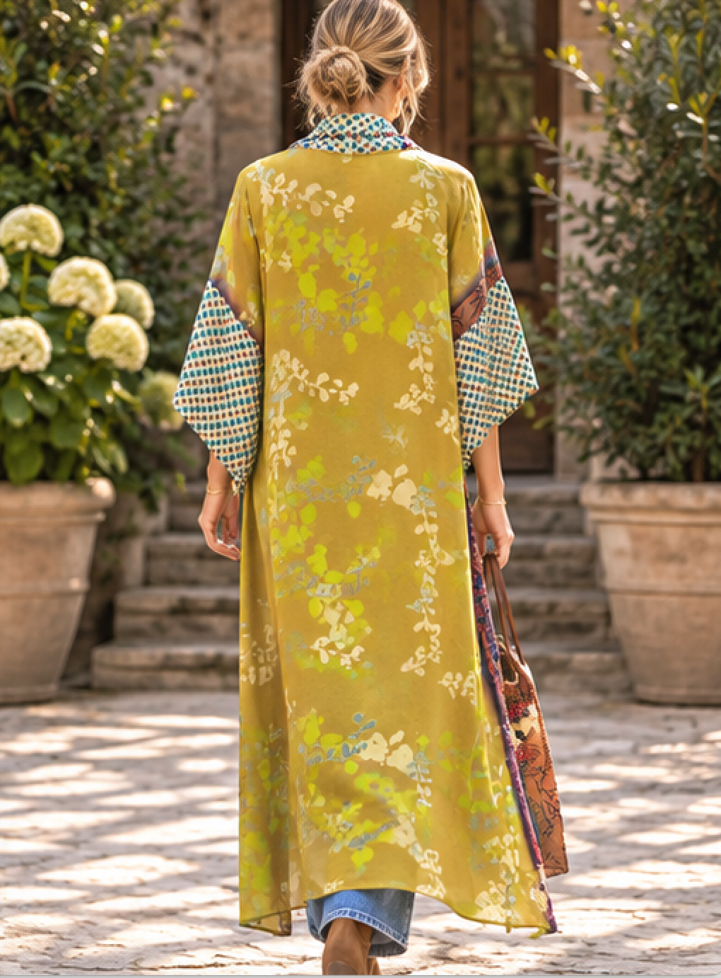 A woman viewed from behind wearing a long olive-yellow kaftan dress with a green and yellow floral pattern, patterned sleeve cuffs, and a blue-and-white checkered collar, walking outdoors in a garden courtyard.
