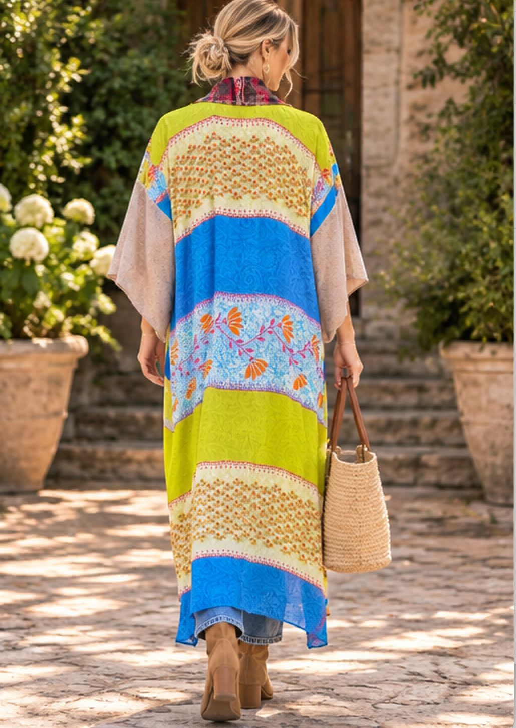 A woman viewed from behind wearing a long, loose kaftan-style dress with a vibrant multicolor pattern of lime green, sky blue, beige, and orange floral motifs. She is walking on a stone path in an outdoor garden setting, holding a woven straw bag.