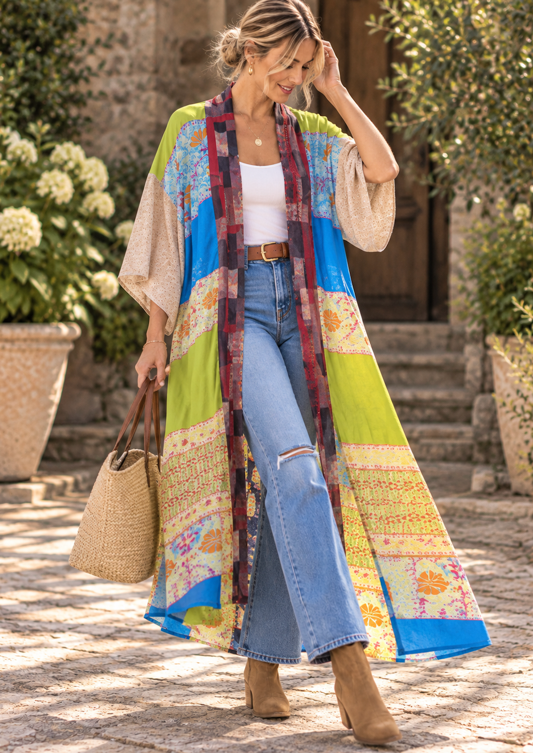 A woman stands outdoors in a stone courtyard wearing a long, loose patchwork kimono-style cardigan with bright blue, lime green, beige, and floral print panels over a white tank top and light blue jeans. She is carrying a woven straw tote bag.