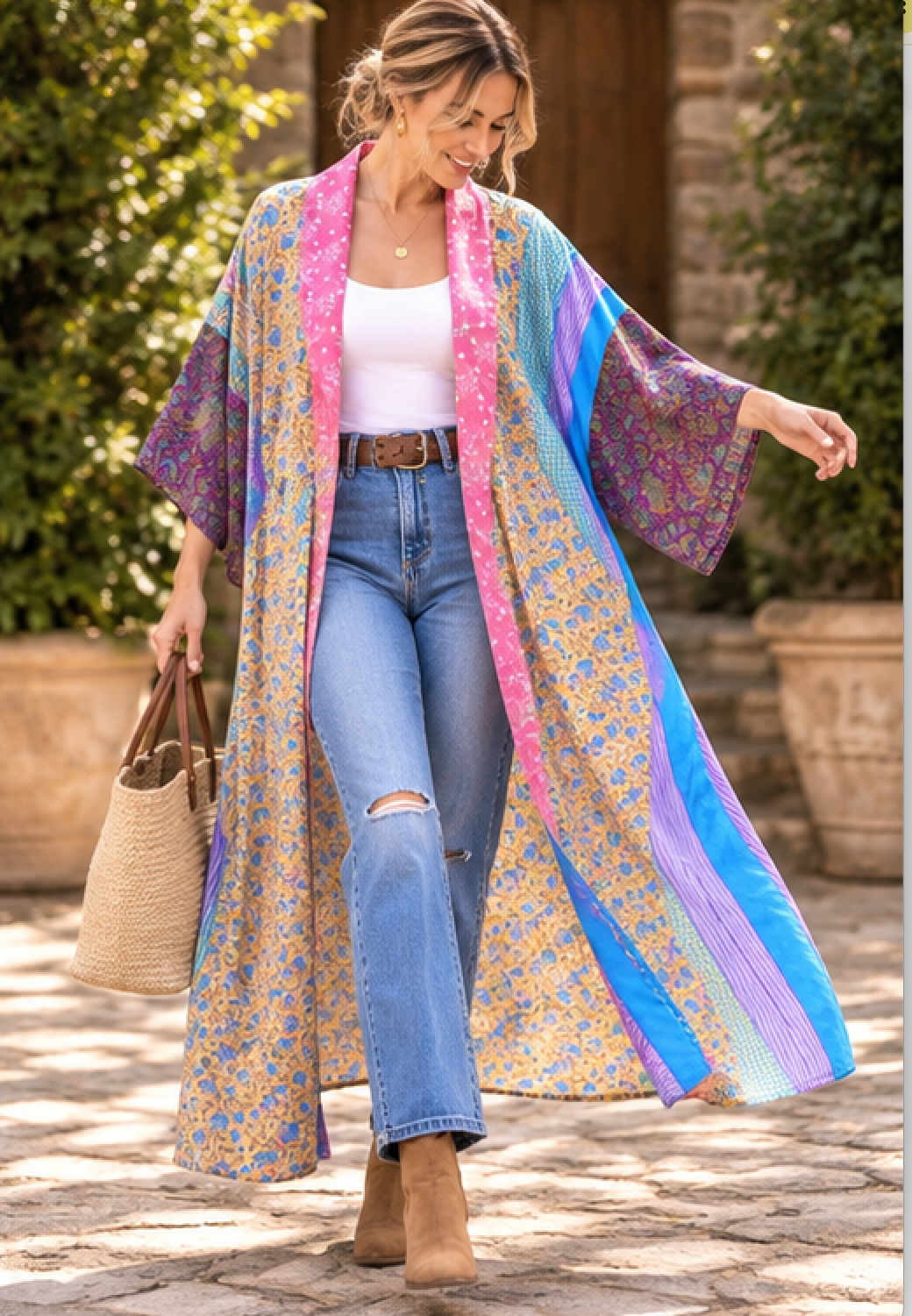 A woman walking outdoors wearing a long, colorful floral kimono-style cardigan with wide sleeves over a white tank top and blue jeans, holding a woven tote bag.