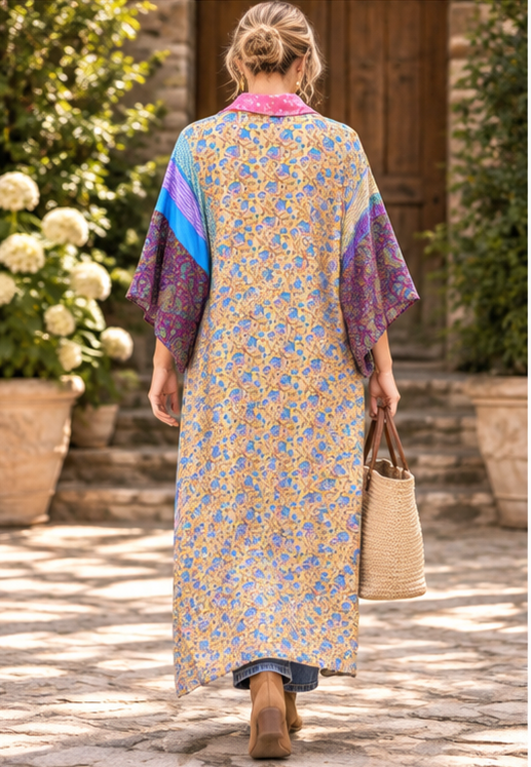 A woman viewed from behind wearing a long, loose kaftan dress with a yellow floral print, purple and blue patterned sleeves, and a pink collar, walking outdoors in a garden setting while carrying a woven straw bag.