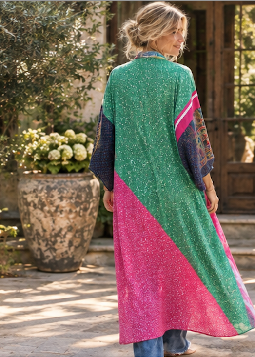 A woman stands on a stone patio in front of a rustic building, viewed from behind as she wears a long, loose patchwork tunic. The tunic features a large green section with subtle gold speckles, a large pink section, and dark navy sleeves with colorful embroidery.