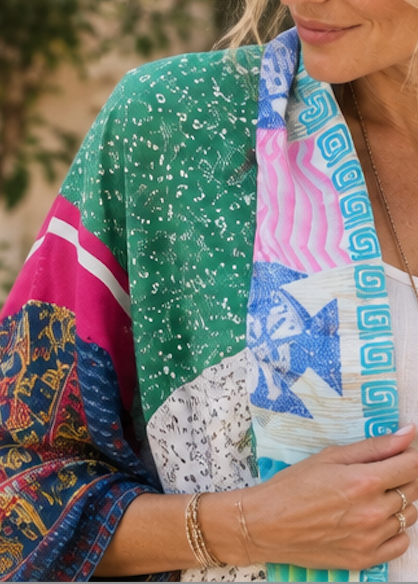 A woman wearing a vibrant, multicolored patchwork kimono cardigan with bohemian patterns, layered over a white tank top and accessorized with gold jewelry.