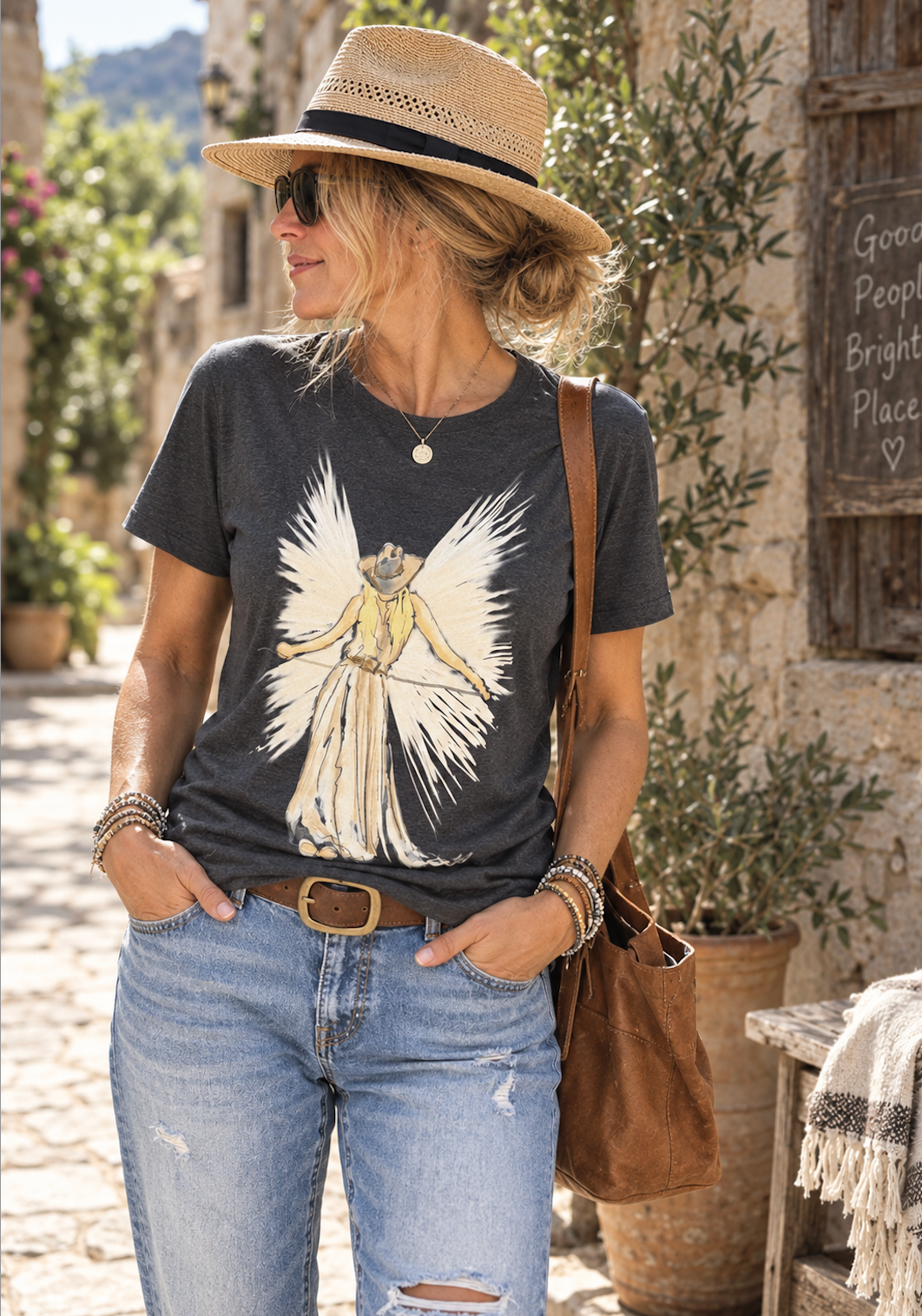 A woman wearing a dark charcoal graphic t-shirt with a cowboy angel illustration, paired with light-wash distressed jeans, a straw hat, and a brown leather shoulder bag, standing in a rustic outdoor setting.