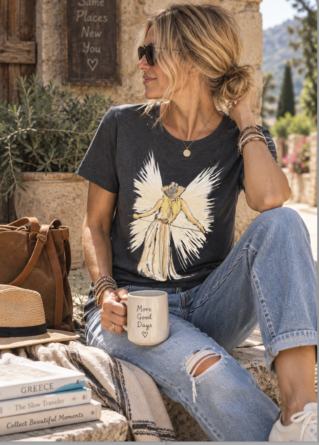 A woman sitting outdoors wearing a dark charcoal graphic t-shirt with a white and yellow angel design, paired with distressed blue jeans and white sneakers. She is holding a white mug with the text 'More Good Days Than Bad'.
