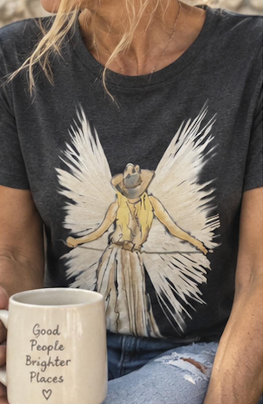 A woman wearing a dark charcoal gray t-shirt with a graphic of an angel with large white wings and a cowboy hat. She is holding a white ceramic mug that reads 'Good People Brighter Places' in black text.